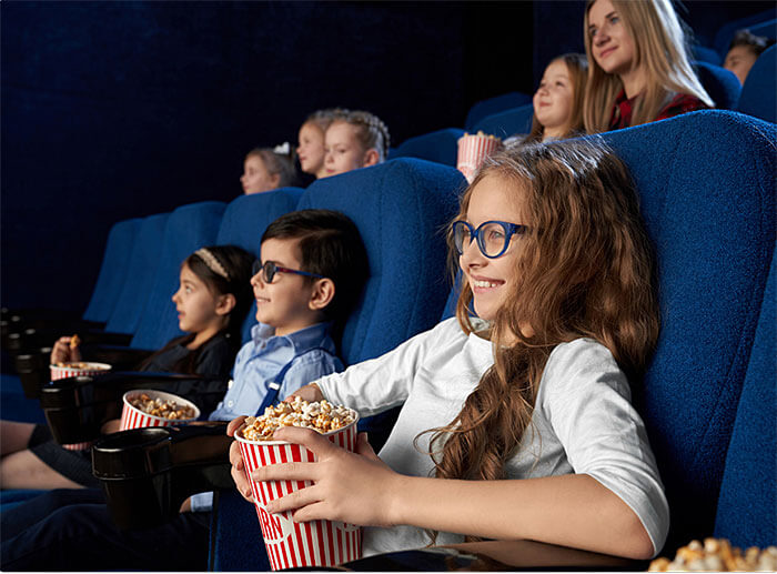 Na zdjęciu widzimy grupę osób w kinie. Są to młode osoby, dzieci. ze skupieniem oglądają coś na ekranie. Na pierwszym planie widać dziewczynkę w okularach, ma duży pojemnik z popcornem.